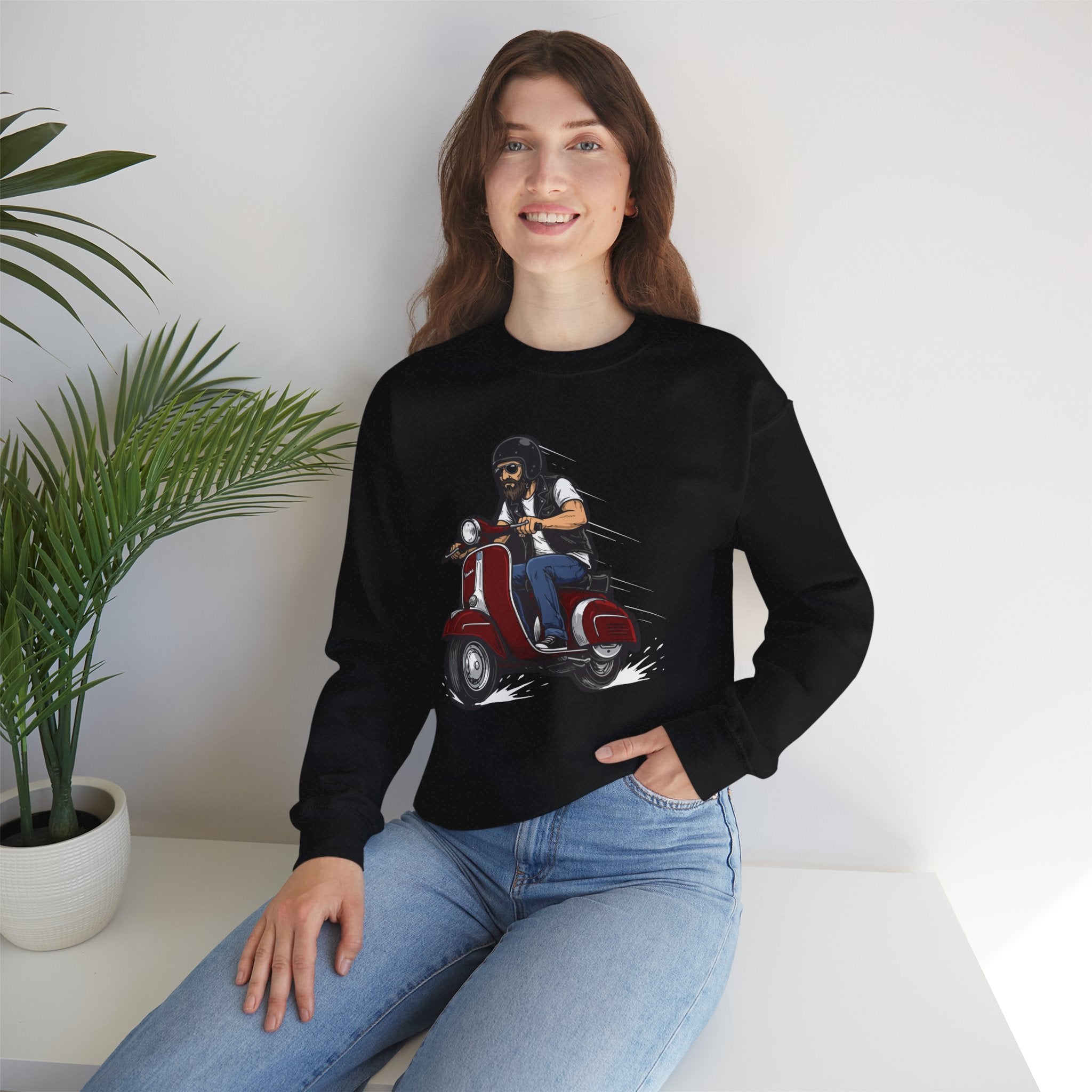 Person wearing a black sweatshirt with a graphic design of a person on a scooter, sitting in a room with a plant.