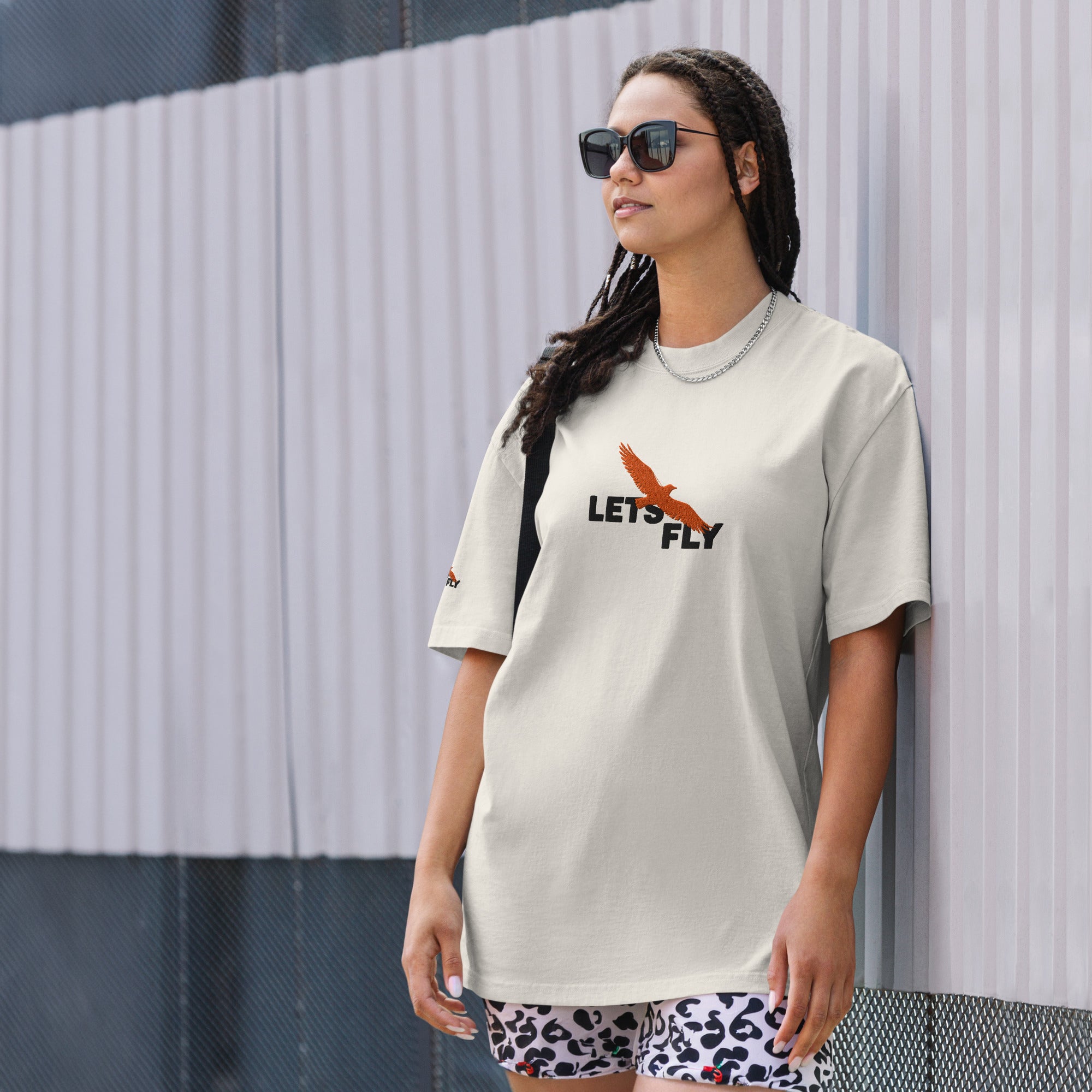 Person wearing a 'Let's Fly' t-shirt with a graphic design against a textured wall.