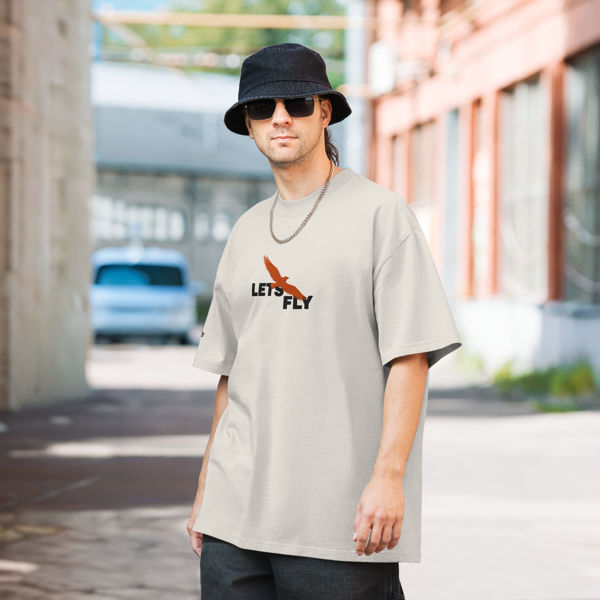 Man wearing a light gray t-shirt with 'Let's Fly' text and graphic, black bucket hat, and sunglasses outdoors.