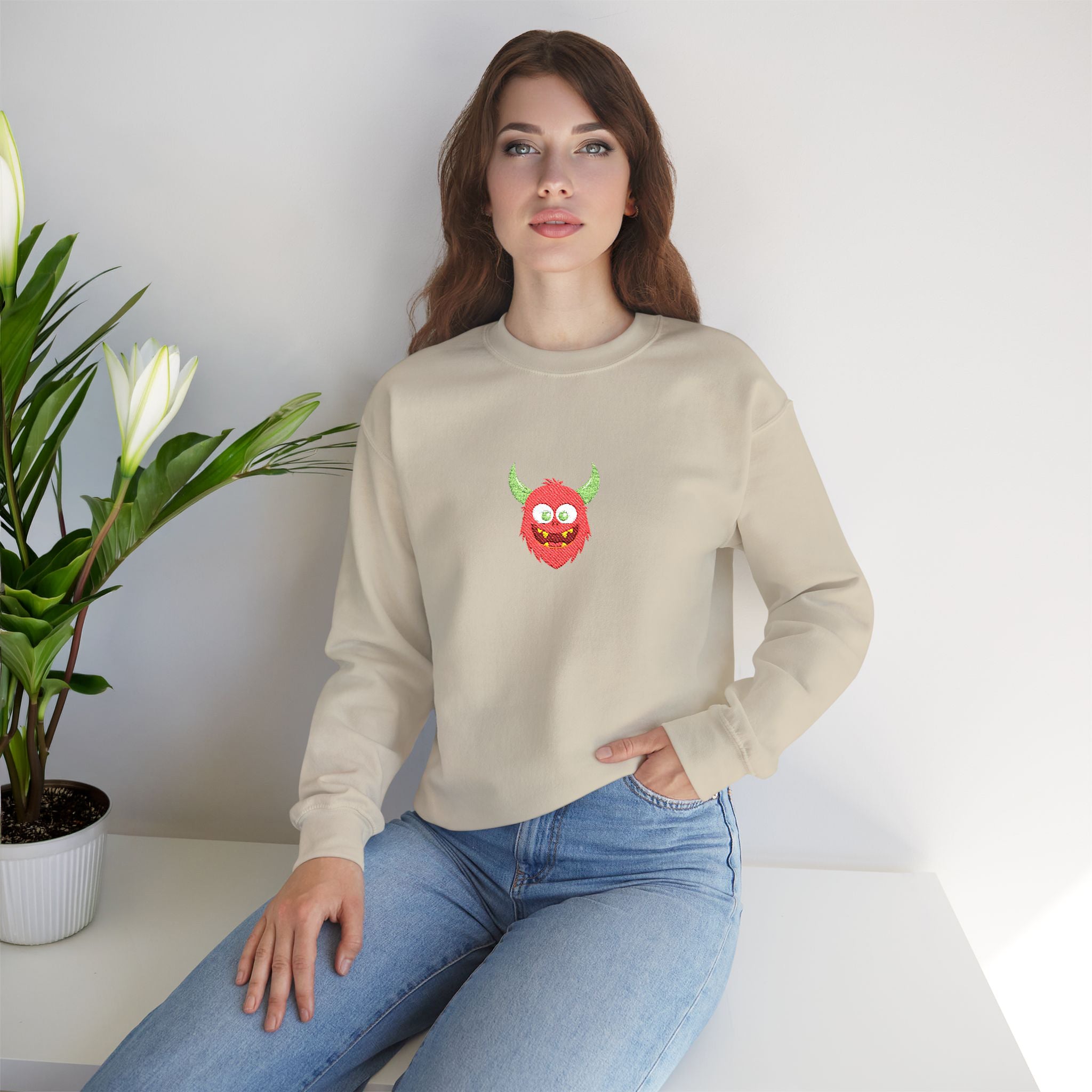 Woman wearing a beige sweatshirt with a colorful graphic design, sitting on a white surface with a plant in the background.