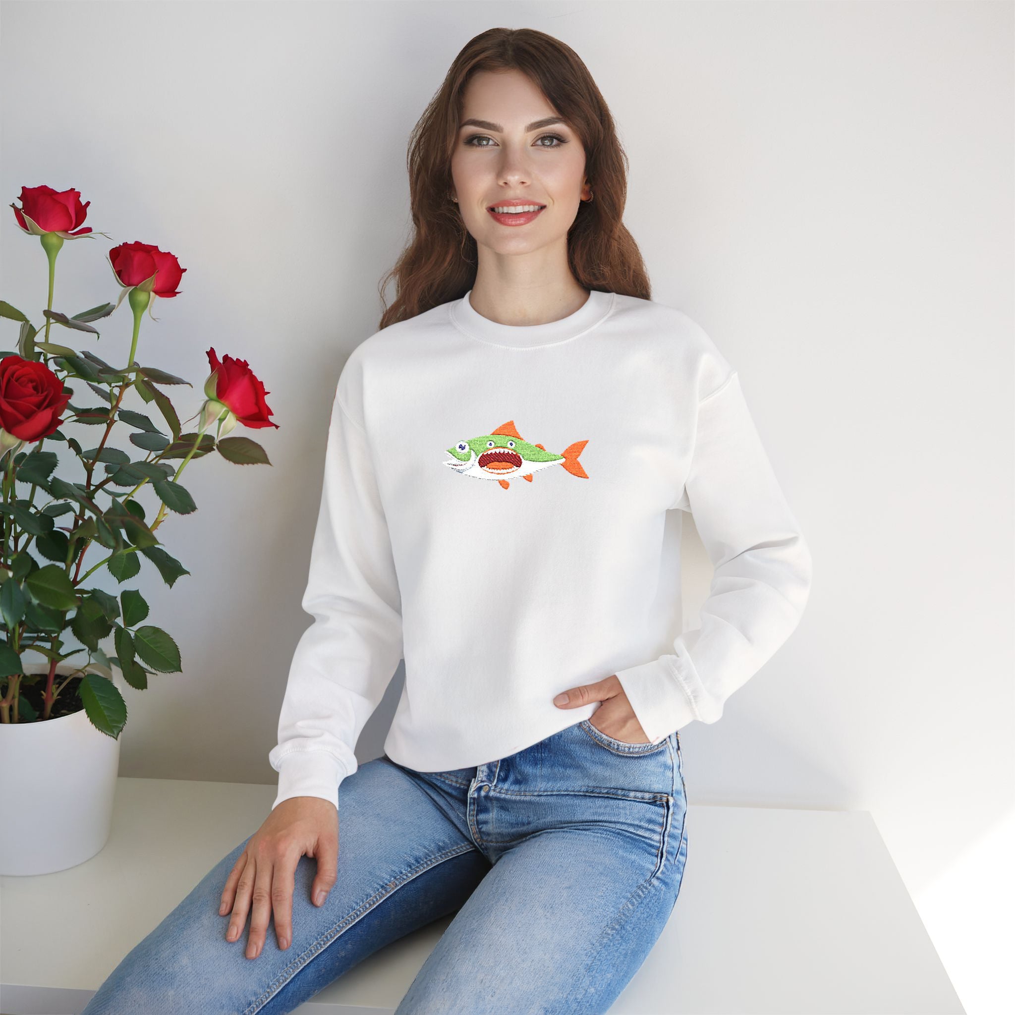Woman wearing a white sweatshirt with a colorful fish design, sitting next to a potted plant with red flowers.