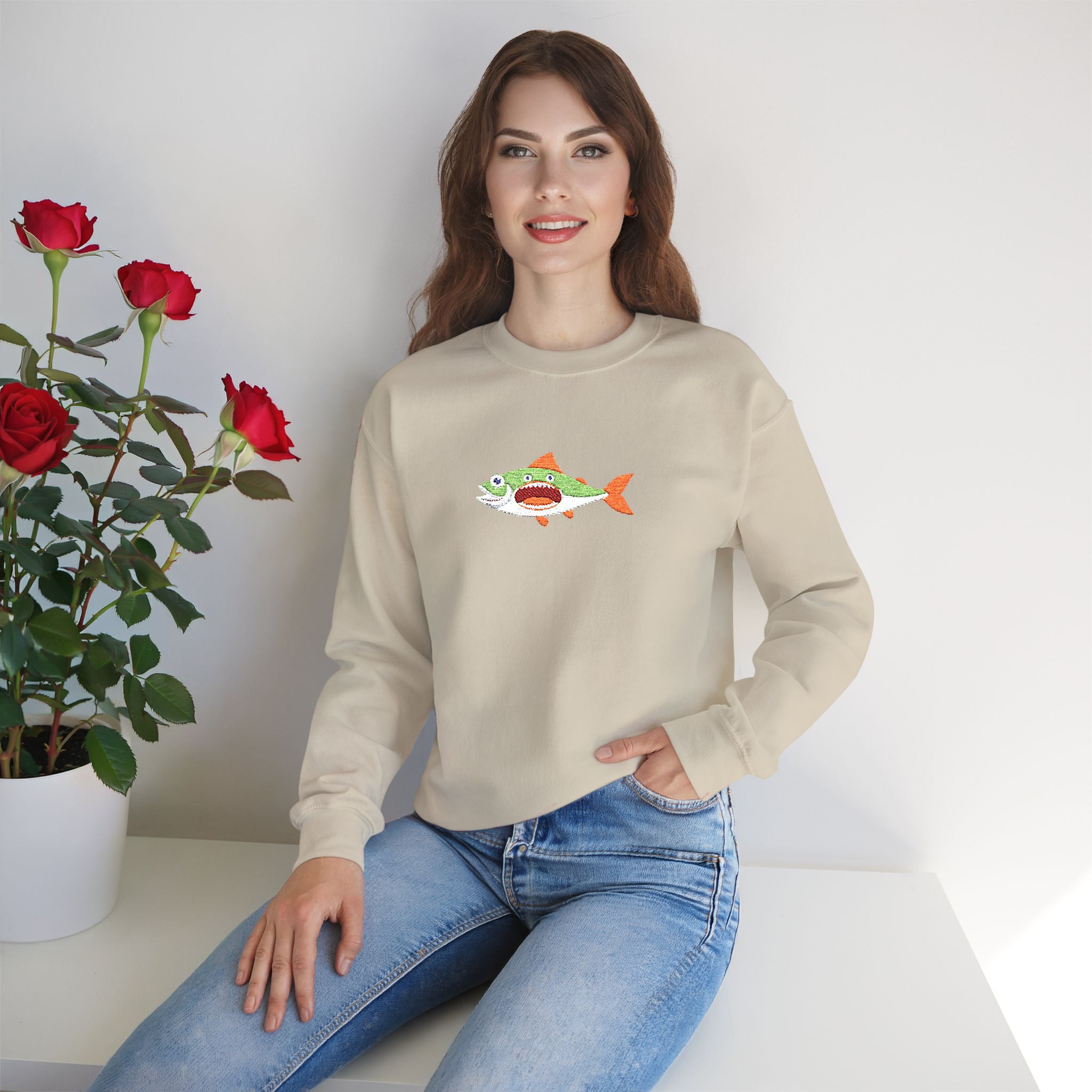 Woman wearing a beige sweatshirt with a fish graphic, sitting on a white surface with red roses and a plant in the background.