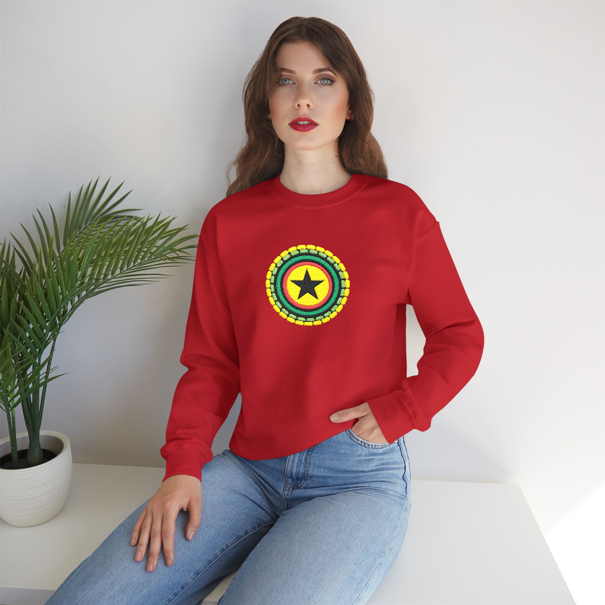Woman wearing a red sweatshirt with a colorful emblem sitting on a white surface with a plant in the background.
